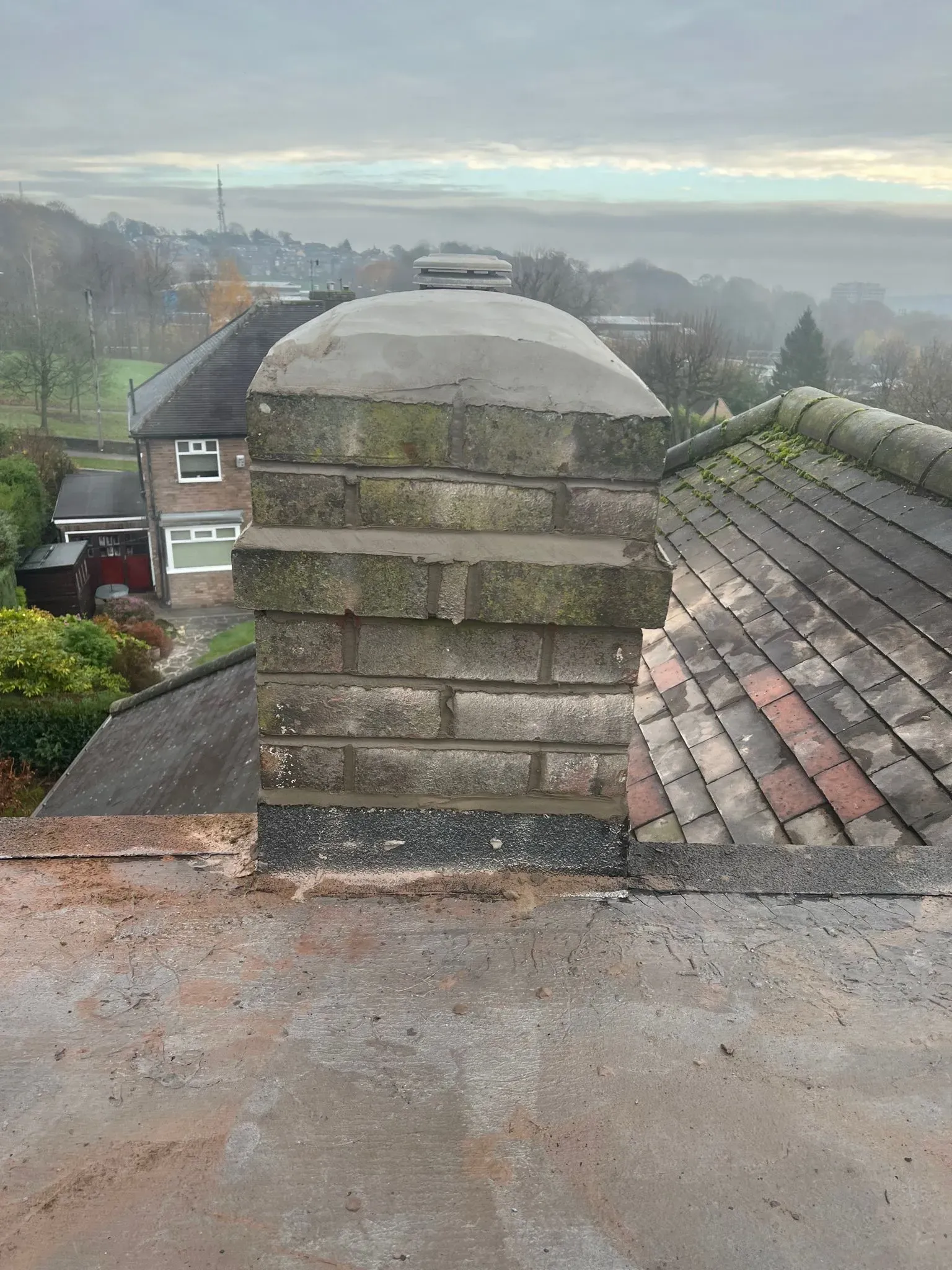 Close-up of a brick chimney on a tiled roof with a view of houses in the background.