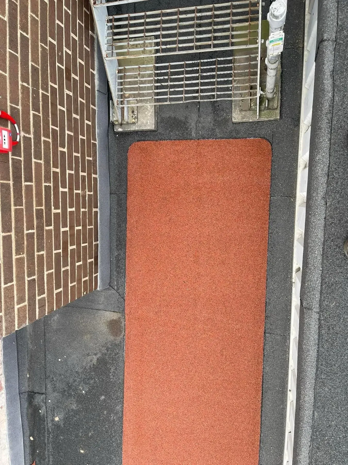 Overhead view of a flat roof area with a red mat, metal grate, and brick wall.