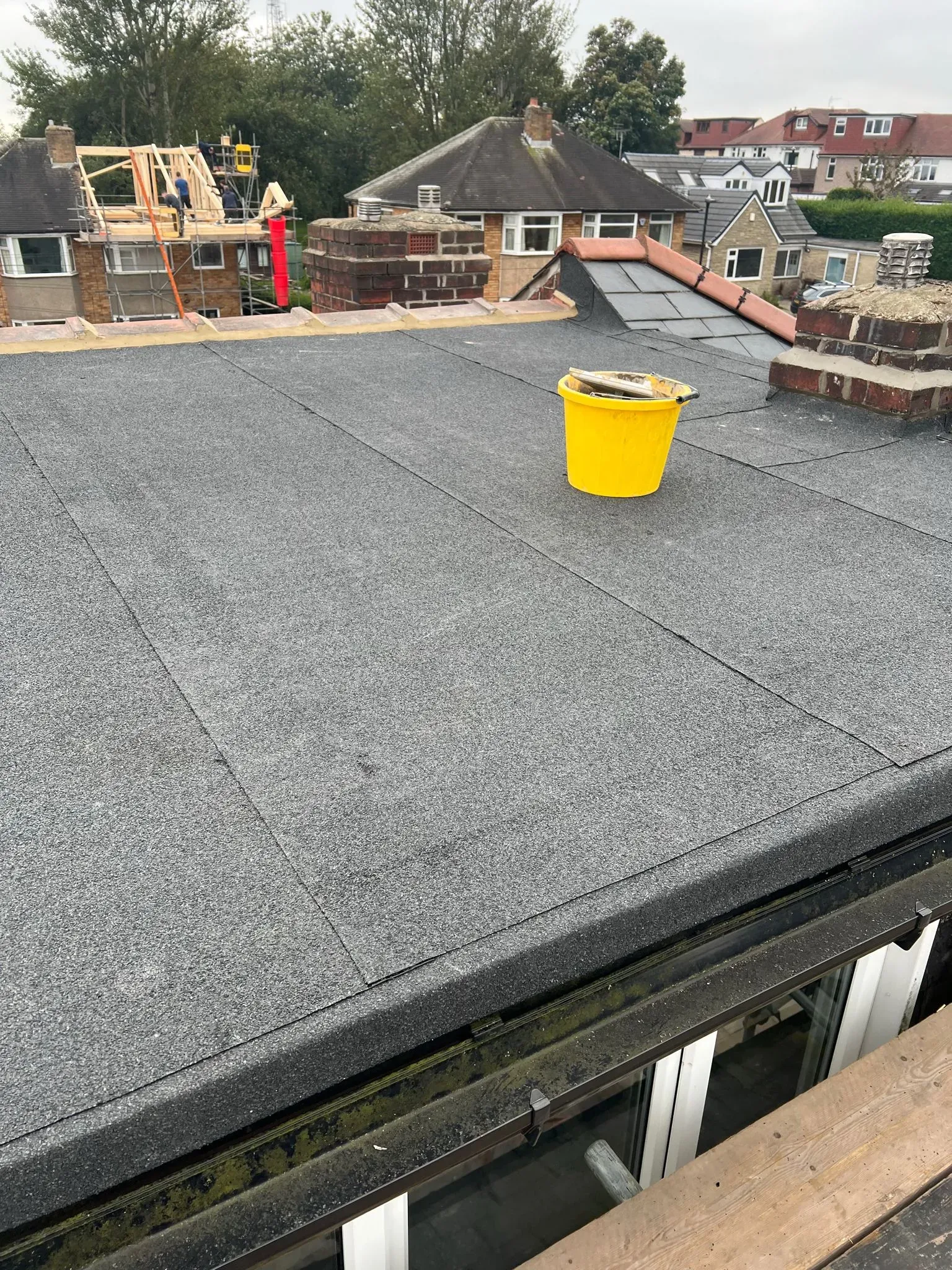 Flat roof surface with a yellow bucket and view of residential houses in the background, one with a new pitched roof under construction.