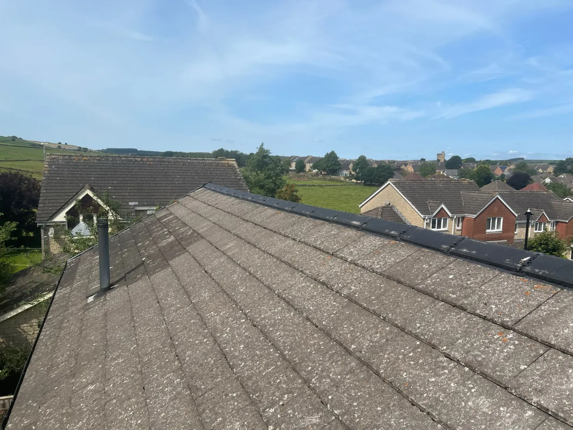 A pitched roof with weathered grey tiles overlooking houses and fields