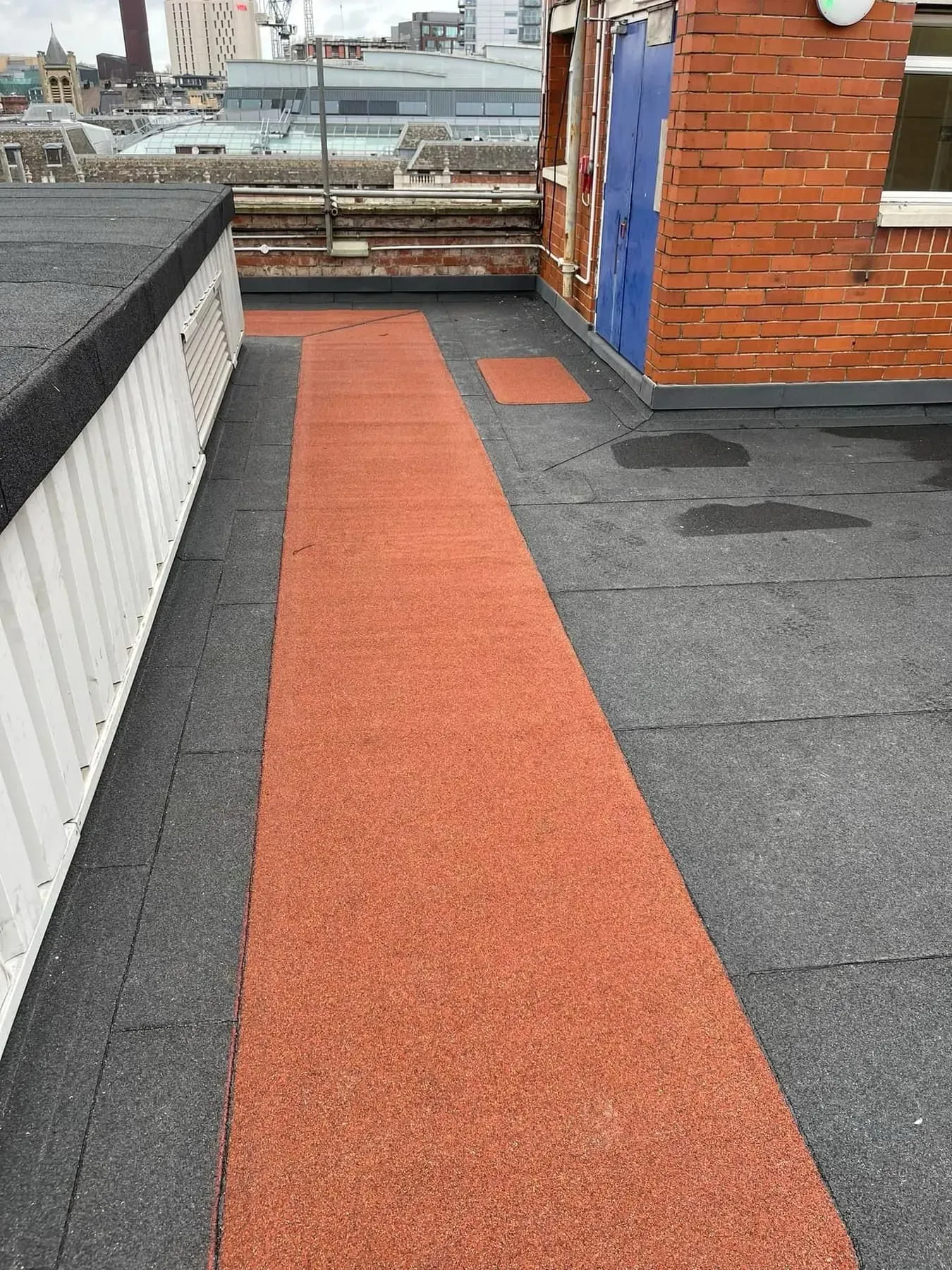 Flat roof surface with red non-slip walkway, grey membrane, puddles, brick wall, blue door, and Sheffield city buildings in the background.